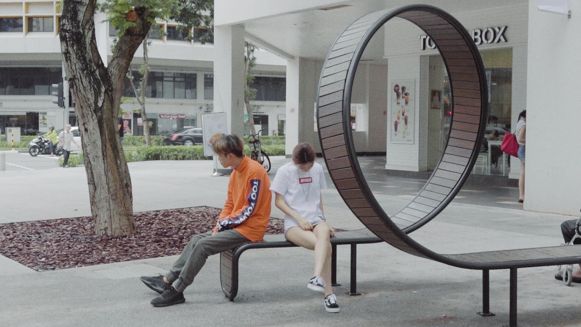 A daylight street scene from SEMENTARA with people gathered around a circular bench.